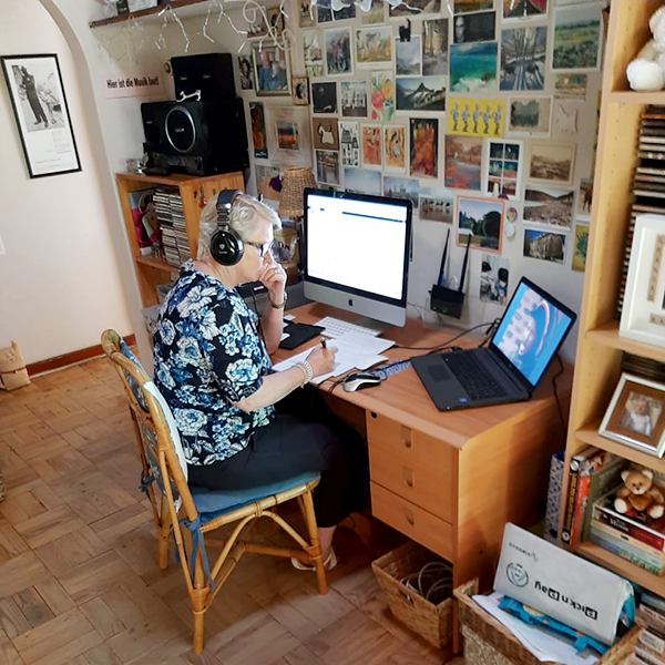 Bev Angus seated at desk against a wall, working on iMac and laptop