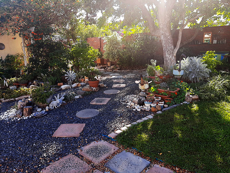 Garden with pebble trail with shrubs, trees and pot plants along each side.