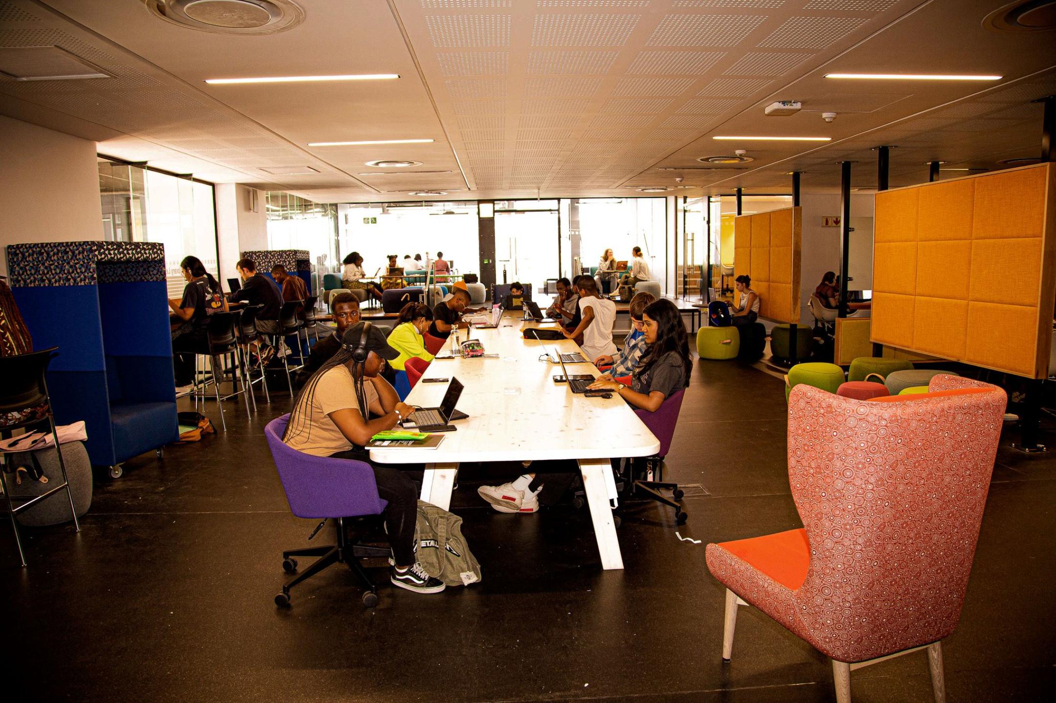 Many students on laptops seated at a long table inside main study space of the Library Learning Lounge, Chris Hani Building