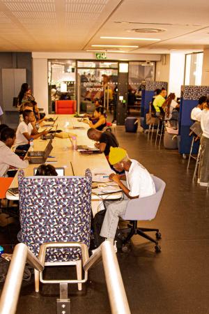 students seated at long table and along window seats on the main floor of the Library Learning Lounge inside Chris Hani Building. Students walking and chattin